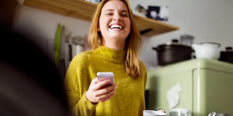 En smilende kvinde iført en gul trøje står i et køkken med en smartphone i hånden og en kop i den anden.