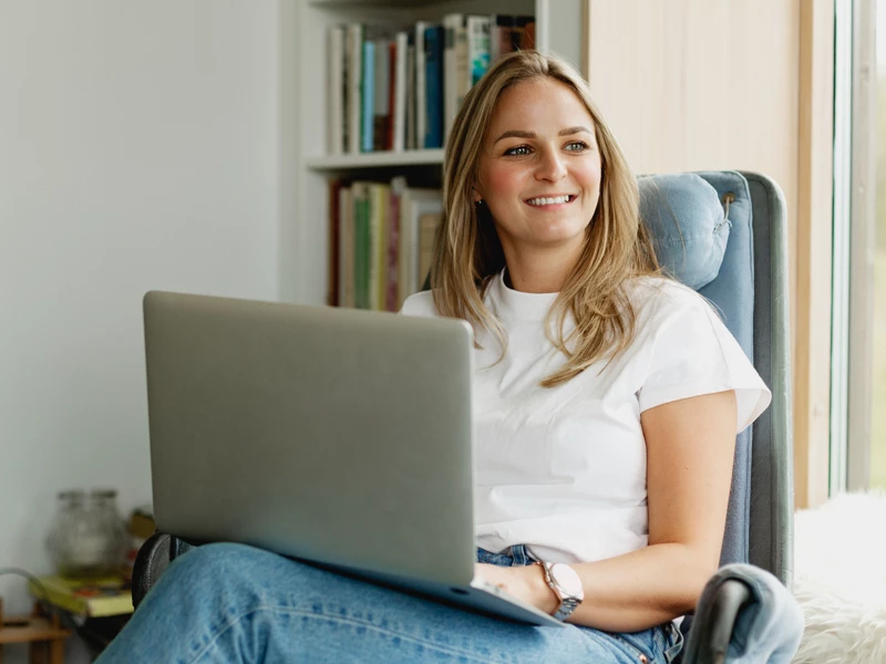 En smilende kvinde sidder afslappet i en lænestol med en bærbar computer på skødet.