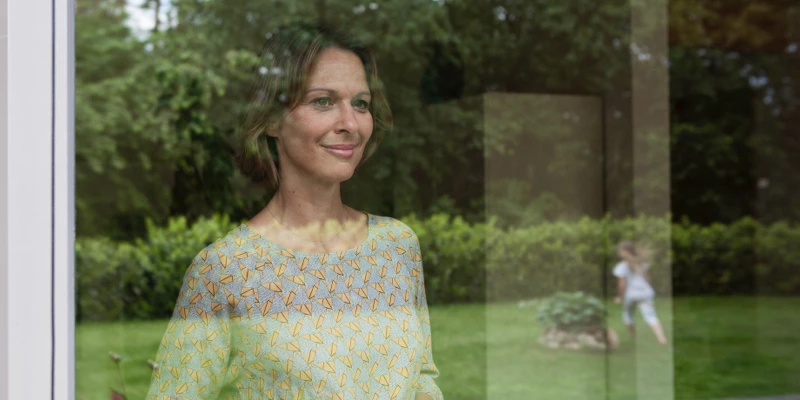 Kvinde ser på sit barn lege i haven fra vinduet