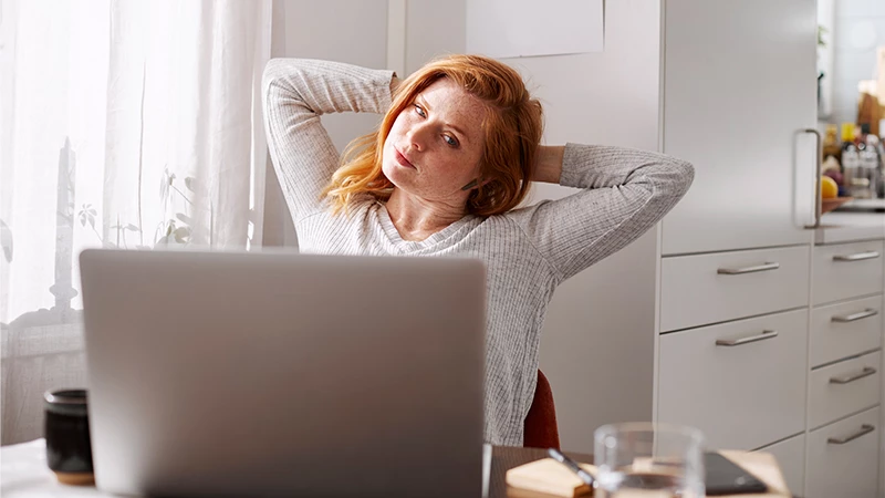 Rødhåret kvinde i køkken foran laptop mens hun strækker sig