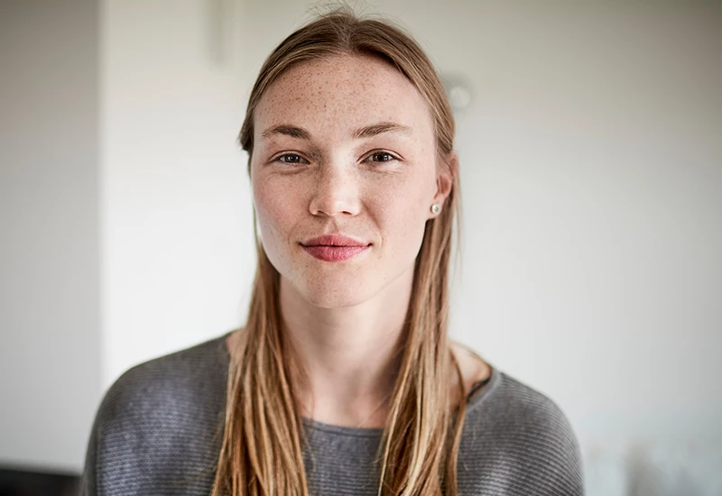 Ung kvinde indenfor med grå trøje på, der kigger i kameraet