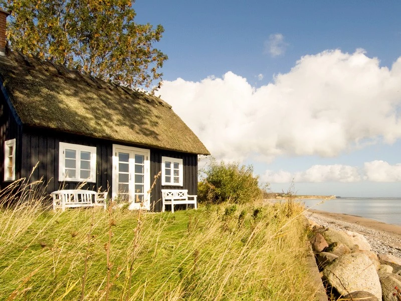 sort sommerhus set udefra med langt græs i forgrund og strand i baggrund